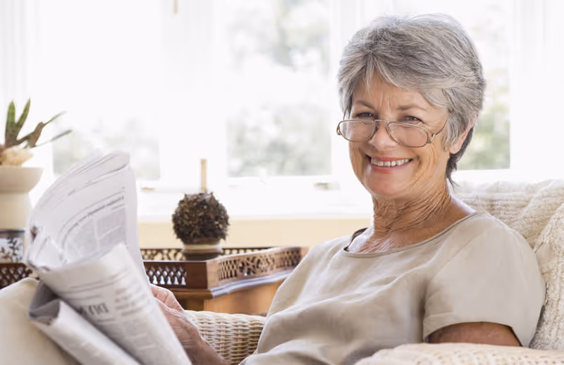 An elderly woman with short gray hair and glasses is sitting comfortably on a cushioned chair, smiling while holding a newspaper. The background shows a bright window and some decorative items on a wooden table.