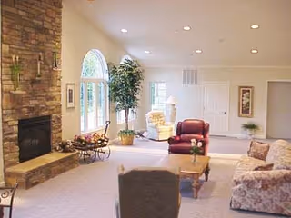 A spacious living room with a stone fireplace on the left wall, a large arched window, and several pieces of comfortable seating including a floral-patterned sofa, a red armchair, and a beige recliner. There is a wooden coffee table in the center and potted plants near the window and door. The room has recessed lighting and light-colored walls and carpet.