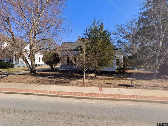 Front exterior of a single-story house with a porch, trees, and a sidewalk under a clear blue sky.