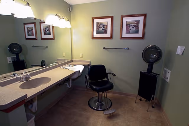 Interior view of a room with a long countertop and sink on the left side, a large mirror above the countertop, a black salon chair in the center, and a vintage hair dryer on a stand to the right. The walls are painted light green and decorated with two framed pictures.