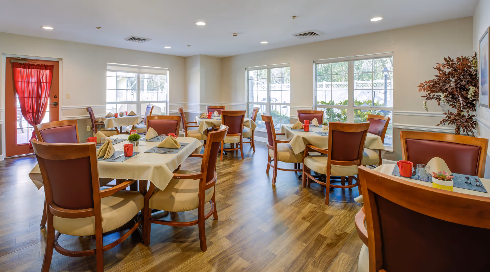A bright dining room with several tables covered with beige tablecloths, each set with folded napkins, red mugs, and small decorative plants. The room has wooden chairs with cushioned seats and backs, large windows letting in natural light, and a red door with a sheer curtain. The floor is wood, and there is a potted plant in the corner.