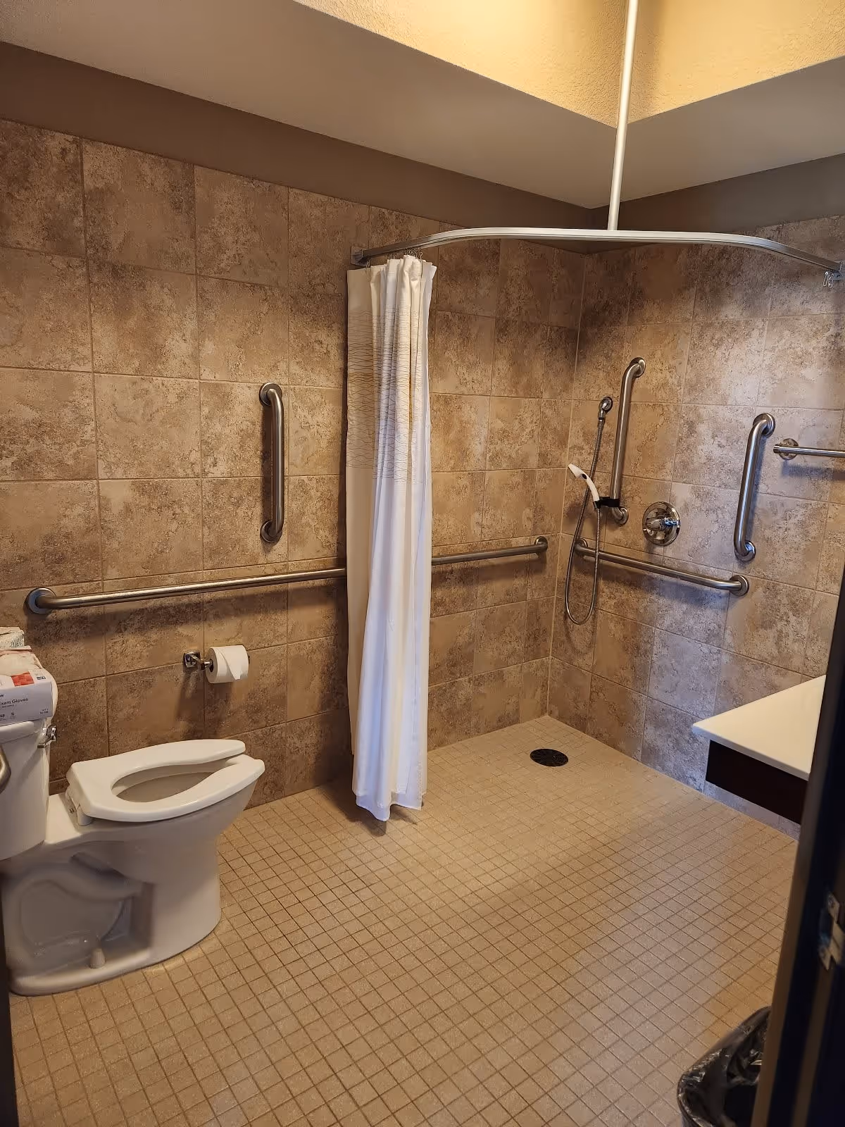 Accessible bathroom with beige tiled walls and floor, featuring a toilet with a raised seat, multiple stainless steel grab bars, a walk-in shower area with a handheld showerhead and a white shower curtain, and a white sink countertop partially visible on the right.
