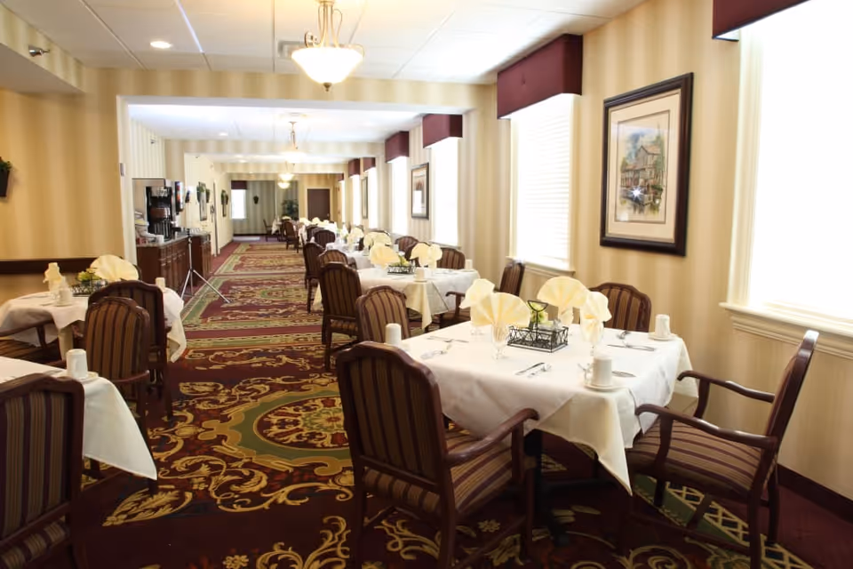 A long dining room with multiple tables covered in white tablecloths, each set with folded napkins, cups, and silverware. The room has patterned carpet, striped upholstered chairs, framed artwork on the walls, and large windows with blinds and valances letting in natural light.
