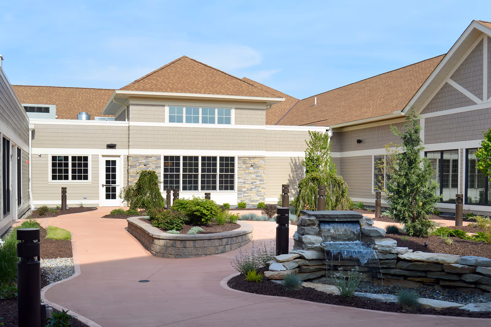 Outdoor courtyard area of a senior living facility with a stone water fountain, landscaped garden beds, small trees, and a paved walkway surrounded by beige buildings with brown shingled roofs under a clear blue sky.