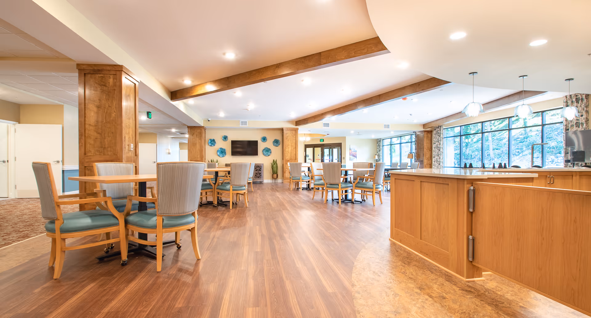 A spacious and well-lit dining area in a senior living facility with wooden floors, multiple tables and chairs arranged for seating, large windows allowing natural light, wooden beams on the ceiling, and a TV mounted on the far wall with decorative plates around it.