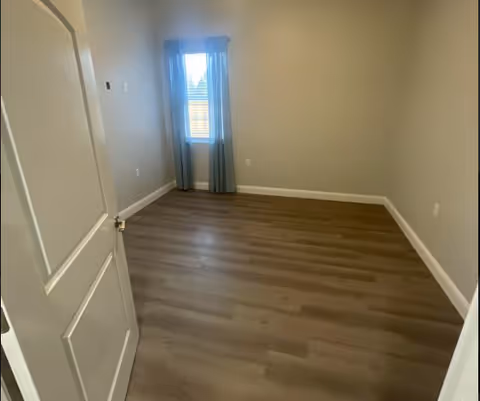 Empty room with light-colored walls, wood flooring, a window with blue curtains, and a partially open white door.