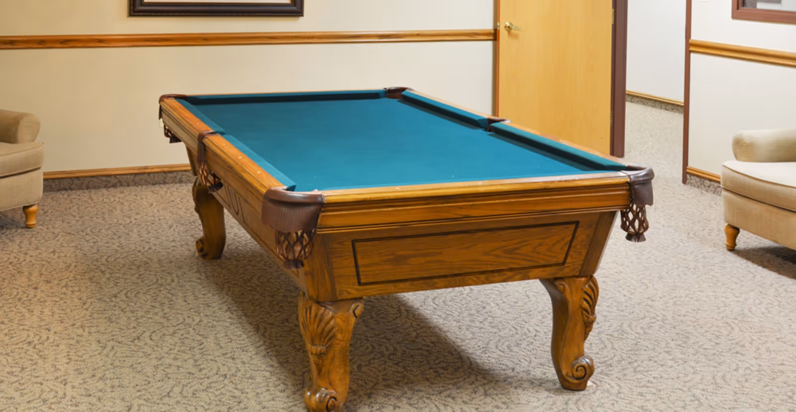 A room with a wooden pool table with green felt top, surrounded by beige carpet and two beige armchairs, with a closed wooden door in the background.