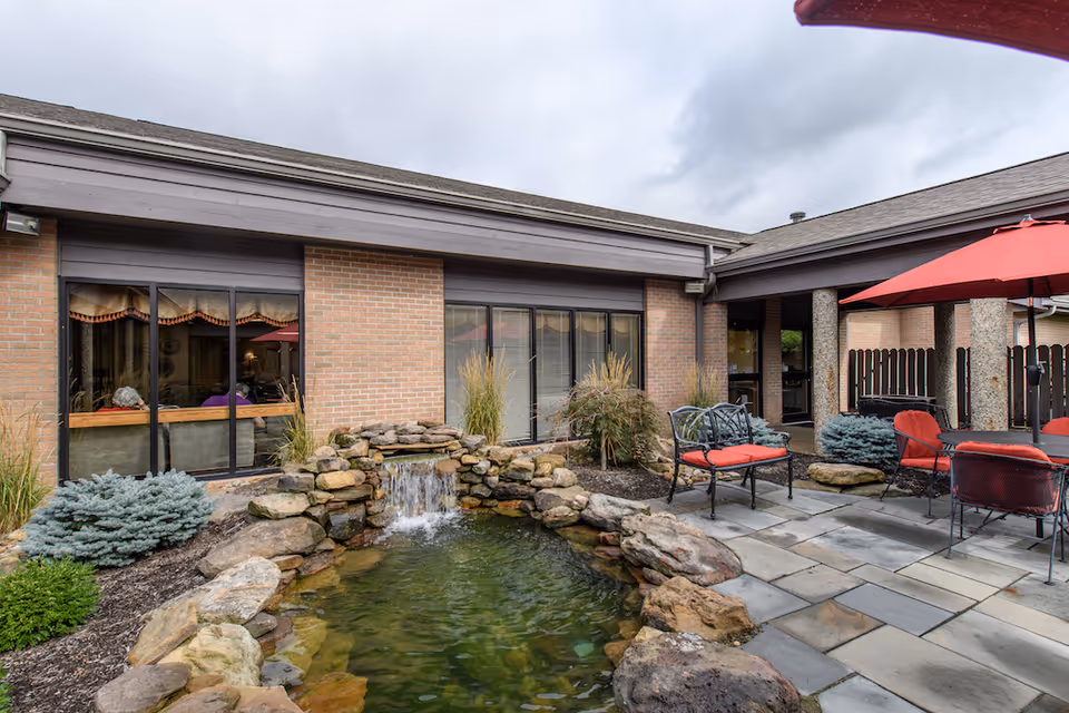 Courtyard patio with a small rock waterfall pond, outdoor seating with red-cushioned chairs and umbrellas in front of a brick building.