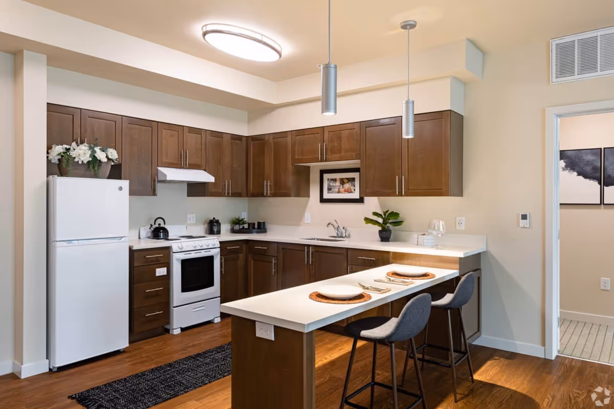 Modern kitchen with an island and two stools, wooden cabinets, white appliances, and pendant lighting.