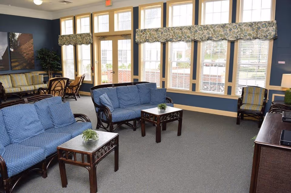 A bright and spacious living room area with large windows letting in natural light. The room features blue cushioned wicker sofas and chairs, two small wooden coffee tables with plants on them, and a patterned valance above the windows. The walls are painted blue with cream trim, and there is a television on a wooden stand to the right.