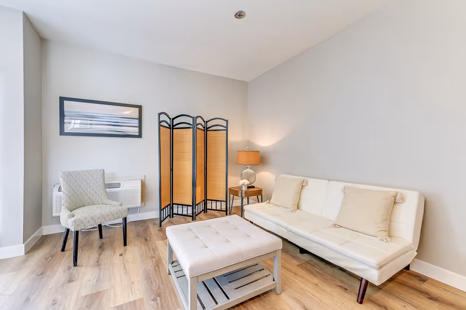 A bright and modern living room with light wood flooring, a white cushioned sofa with two beige pillows, a matching cushioned ottoman, a patterned armchair, a wooden side table with a lamp, and a decorative folding screen against a light gray wall.