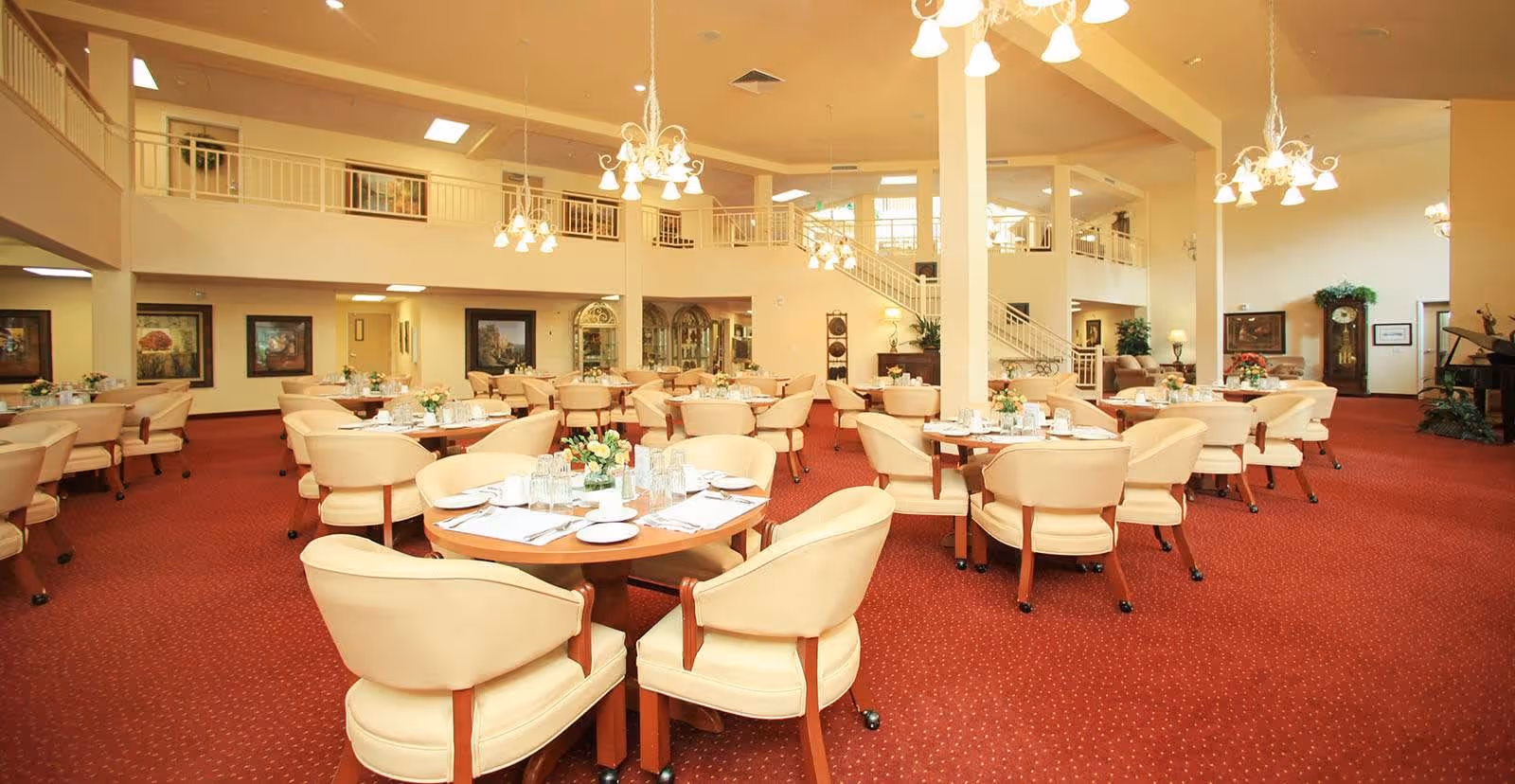 Spacious dining room with multiple round tables set with plates, glasses, and napkins. The room features cream-colored cushioned chairs, red carpet flooring, chandeliers hanging from the ceiling, and a second-floor balcony overlooking the area. There are framed paintings on the walls and a grandfather clock in the corner.