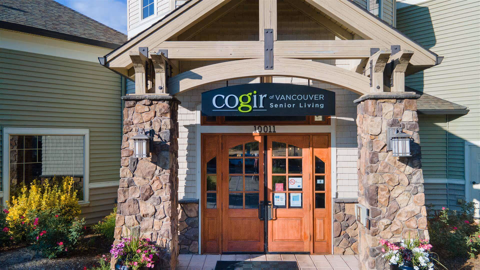 Front entrance of a senior living building with wooden double doors, stone pillars, a gabled porch, and a sign reading "Cogir of Vancouver Senior Living".