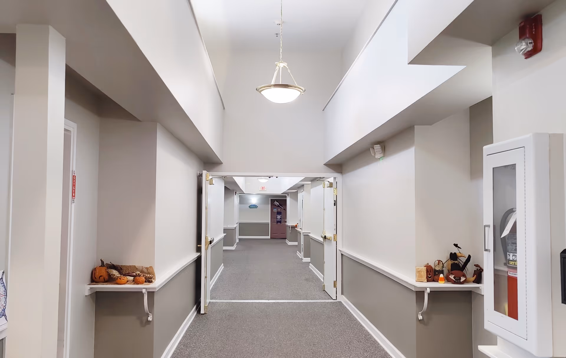 A wide hallway in an assisted living facility with beige and gray walls, carpeted floor, and white double doors open in the middle. Small shelves on either side of the hallway hold autumn-themed decorations including small pumpkins and gourds. A fire extinguisher cabinet is mounted on the right wall, and a hanging light fixture is visible on the ceiling.