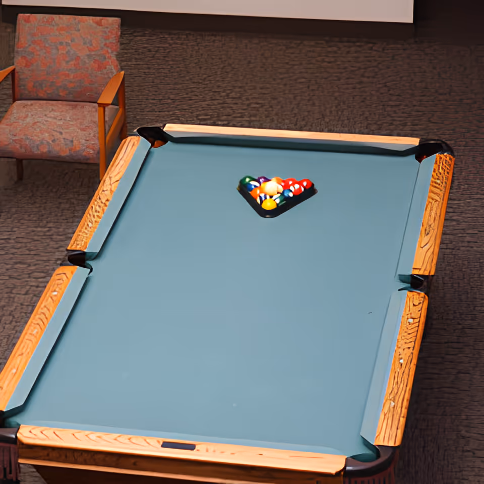A pool table with a green felt surface and a wooden frame, set up with a rack of colorful billiard balls in the center. To the left of the pool table, there is a cushioned armchair with a patterned fabric. The floor is carpeted with a textured design.
