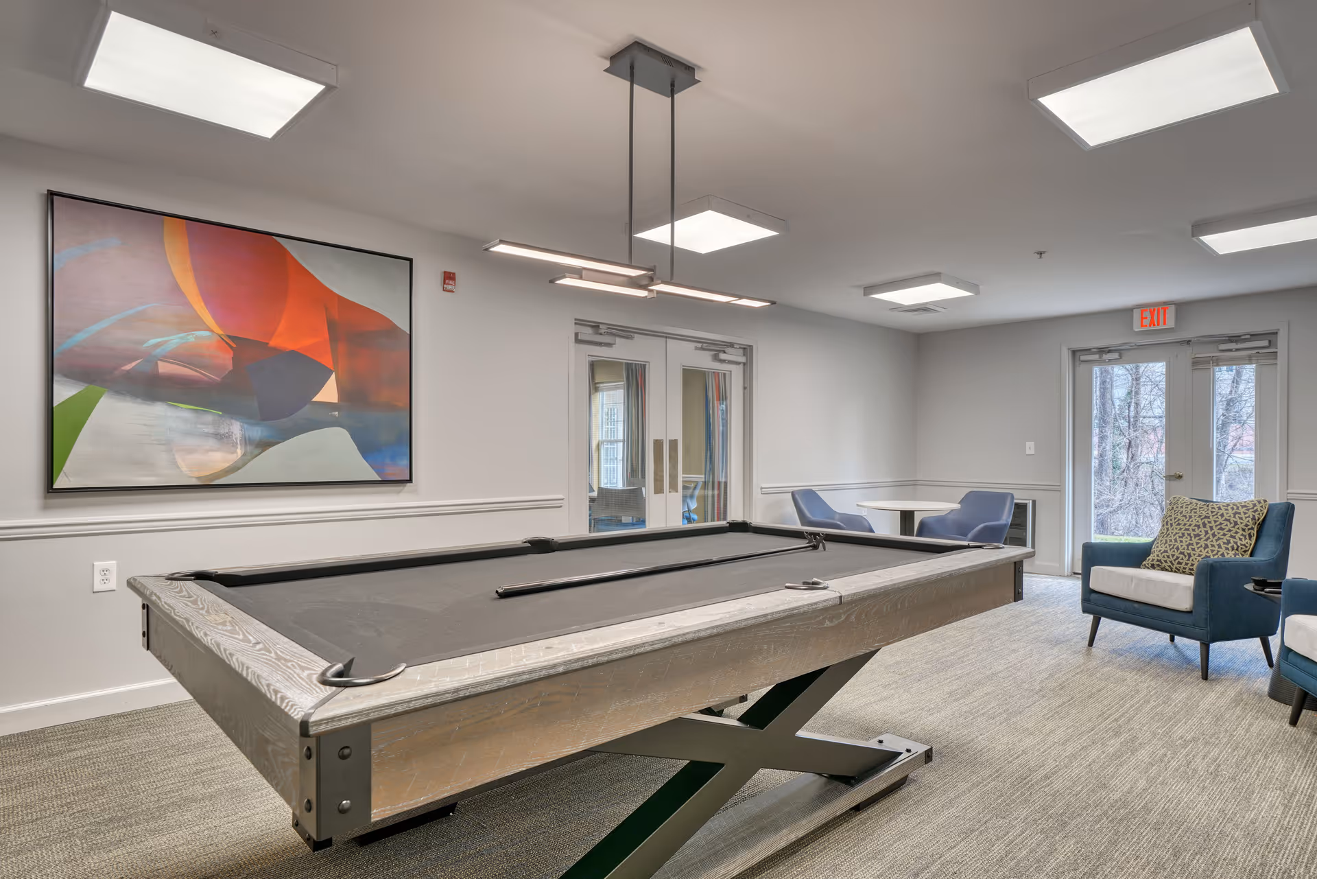 A well-lit recreational room featuring a modern pool table with a black felt surface in the center. The room has light gray walls, a large colorful abstract painting on one wall, and ceiling lights. There are two blue armchairs with patterned cushions near glass doors that lead outside, and a small round table with two blue chairs in the corner.