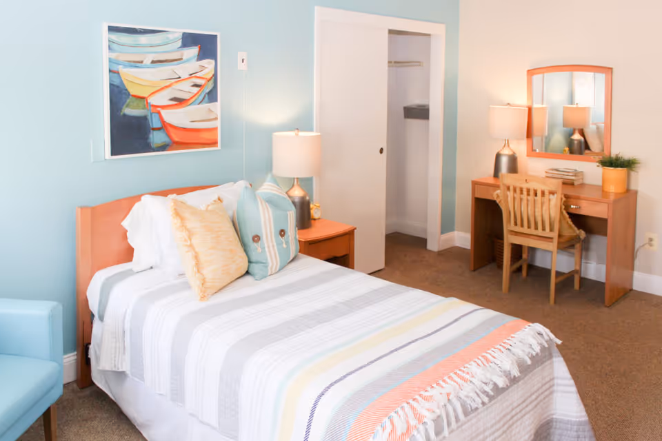 A bright and cozy bedroom with a single bed featuring a striped blanket and multiple pillows. There is a wooden nightstand with a lamp and an alarm clock next to the bed. A colorful painting of boats hangs on the light blue wall above the bed. Across from the bed is a wooden desk with a chair, a lamp, books, and a small potted plant. The room has beige carpet and a closet with a sliding door.