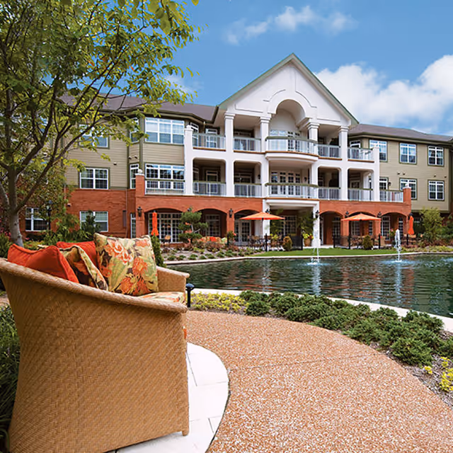 Outdoor seating area with a wicker chair and floral cushions next to a curved pathway, overlooking a pond with fountains in front of a multi-story residential building with balconies and orange umbrellas.