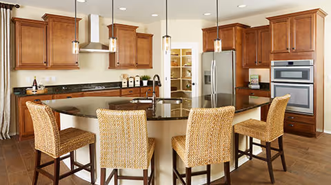 Modern kitchen with wooden cabinets, a large curved island with a dark countertop, four wicker bar stools, stainless steel refrigerator, built-in oven, and pendant lights hanging from the ceiling.