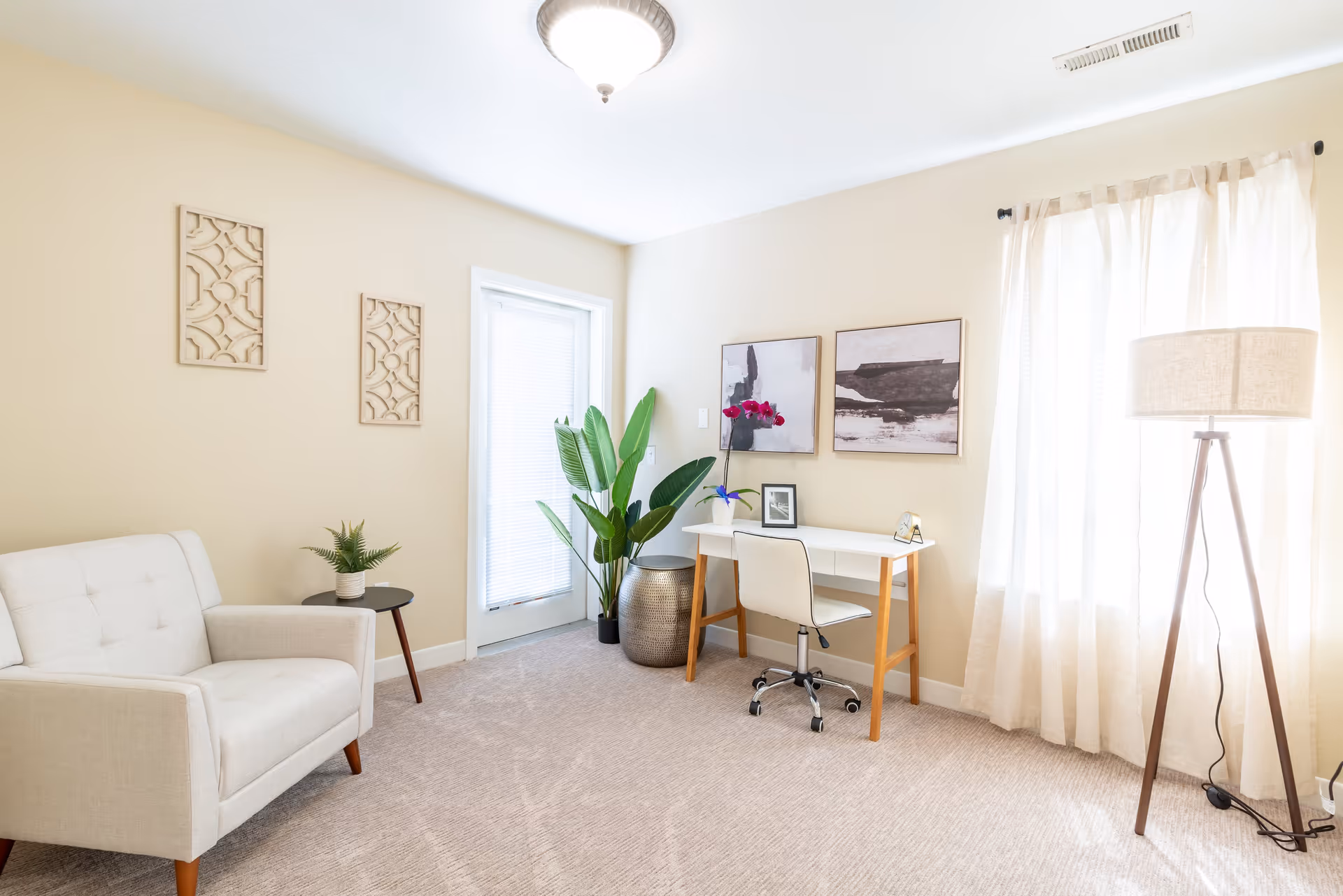 A bright and airy room with beige walls and carpeted floor featuring a white cushioned armchair next to a small round side table with a potted plant. A white desk with wooden legs holds a small framed picture, a decorative item, and a vase with pink flowers. Two abstract paintings hang above the desk. A tall green plant in a metallic pot is placed near a door with a window covered by blinds. A large window with sheer curtains allows natural light to fill the room, and a tripod floor lamp stands beside it.