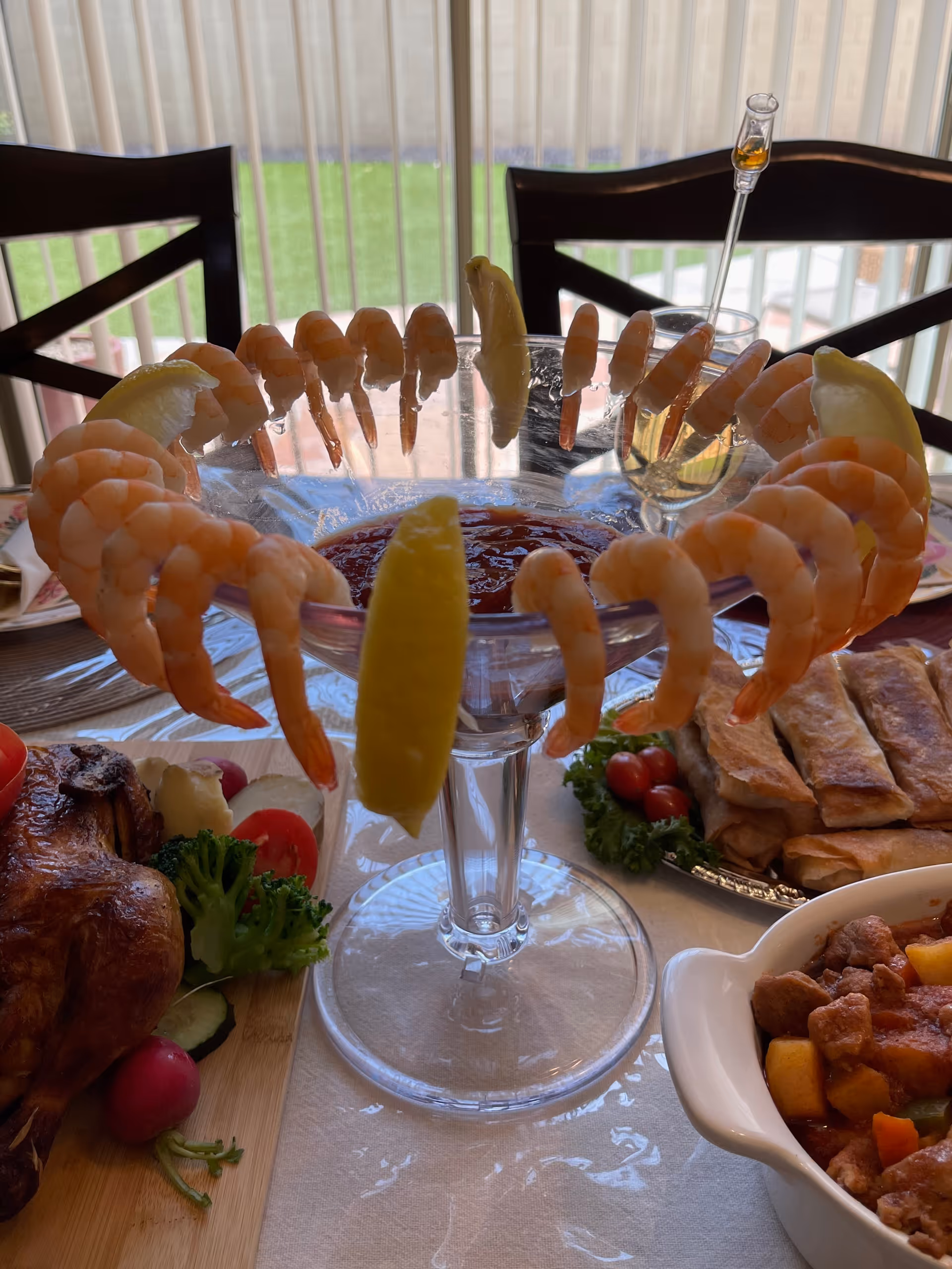A glass shrimp cocktail centerpiece with lemon wedges on a dining table surrounded by other dishes and chairs in front of vertical blinds.