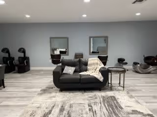 Modern common room with a gray sofa and side table on a patterned rug, with two mirrors and salon-style chairs along a light gray wall.