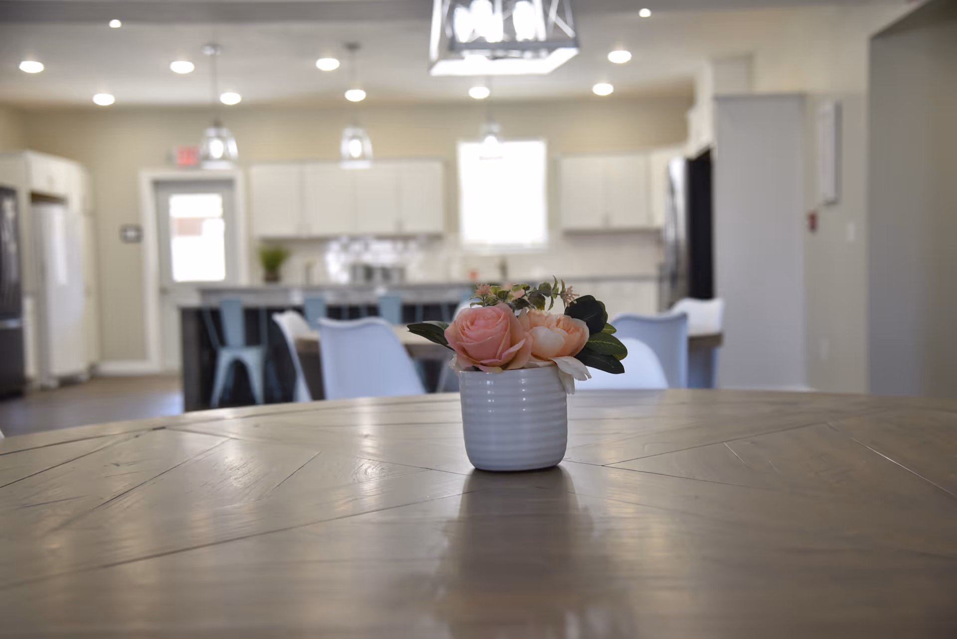 Small vase of pink flowers centered on a wooden table with an open kitchen and dining area blurred in the background.