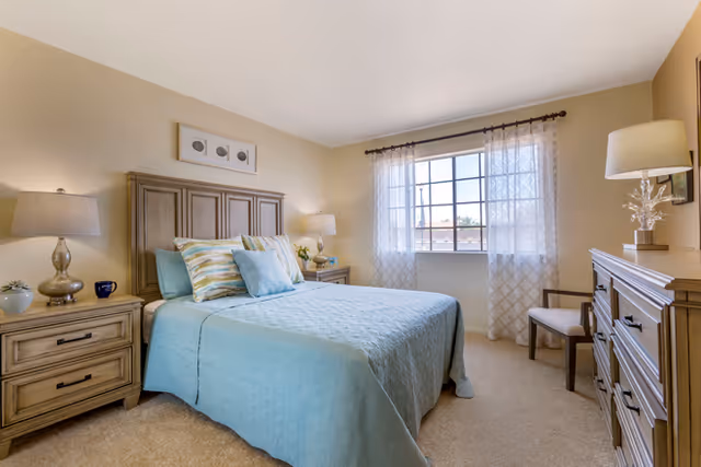 A bright bedroom with a large window covered by sheer white curtains. The room features a wooden bed with a light blue bedspread and multiple pillows, two matching wooden nightstands with lamps, a wooden dresser with a lamp and decorative items, and a chair near the window. The walls are painted beige and the floor is carpeted.
