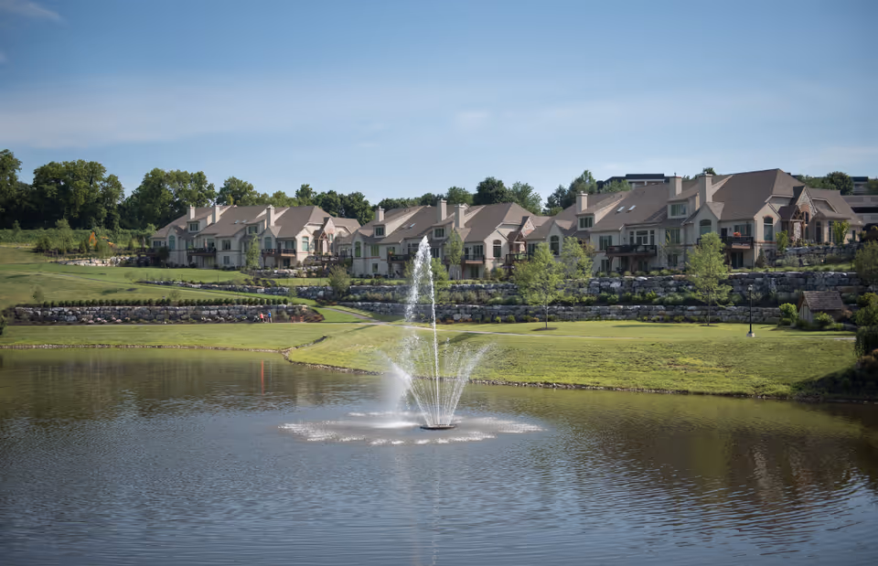 Row of manor-style residences overlooking a pond with a central fountain and terraced lawns.