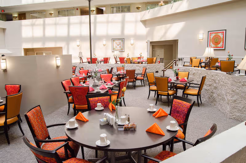 A bright and spacious dining room in a senior living community with multiple round and square tables set with white cups, saucers, and orange napkins. The chairs have colorful upholstery in shades of red, orange, and yellow. The room features a high ceiling with natural light coming through large windows, and framed artwork is displayed on the walls.