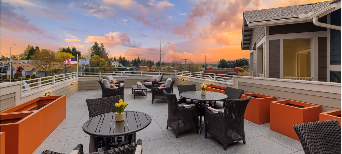 Outdoor patio area at sunset with multiple black wicker chairs and round tables, orange planter boxes along the railing, and an American flag in the background.