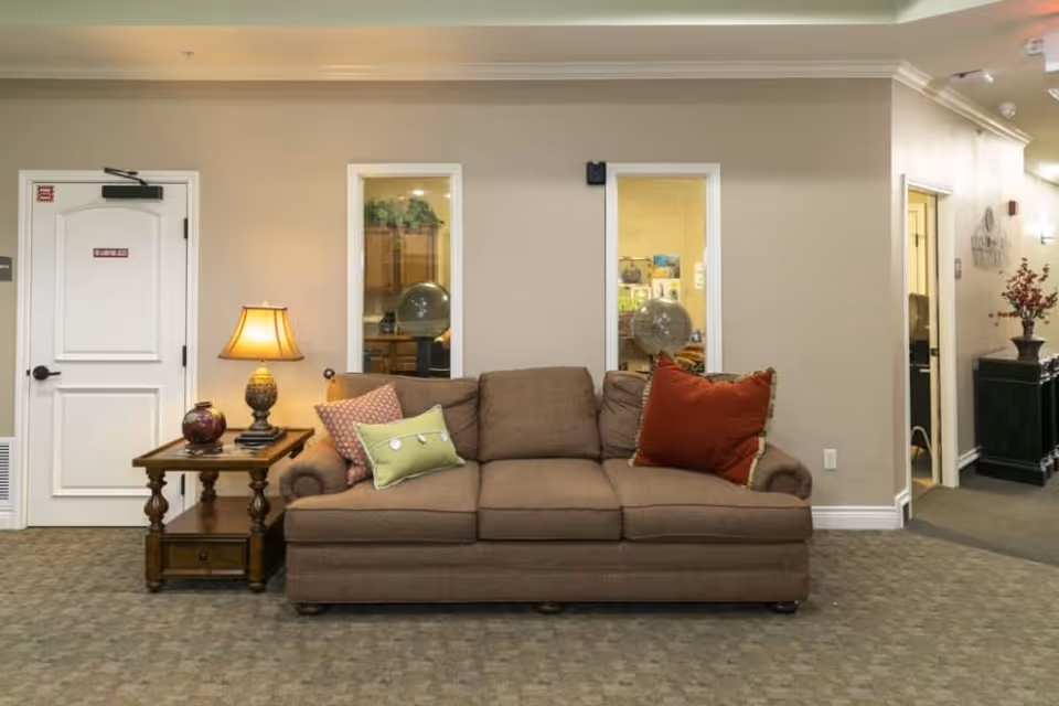 Brown sofa with colorful pillows sits against a beige wall beside a lamp-topped side table and hallway doors in a communal living area.