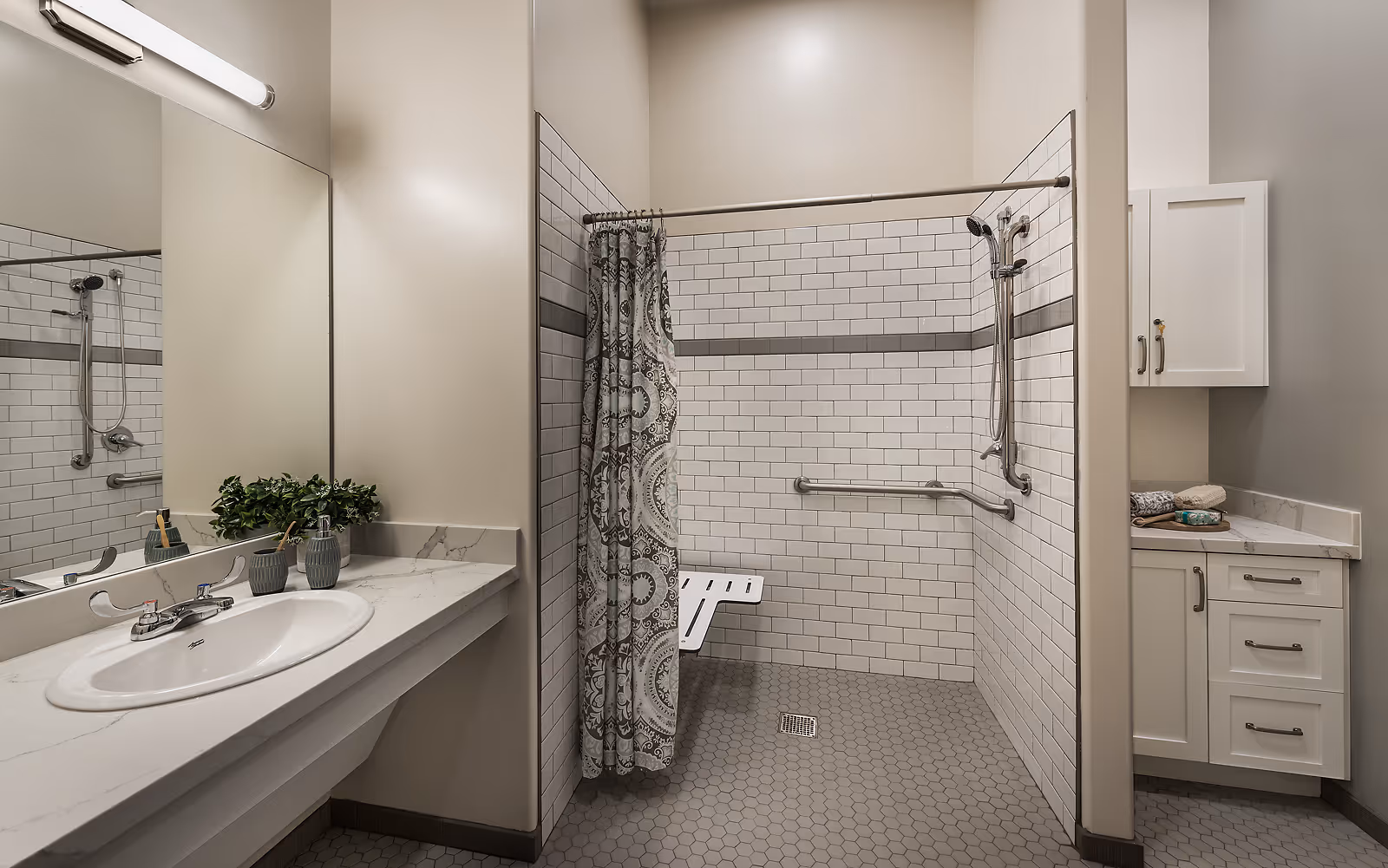 A modern accessible bathroom with a white sink and countertop on the left, a large mirror above the sink, and a walk-in shower with white subway tiles and a patterned shower curtain. The shower has grab bars and a fold-down seat. On the right, there is a white cabinet with drawers and folded towels on the countertop.