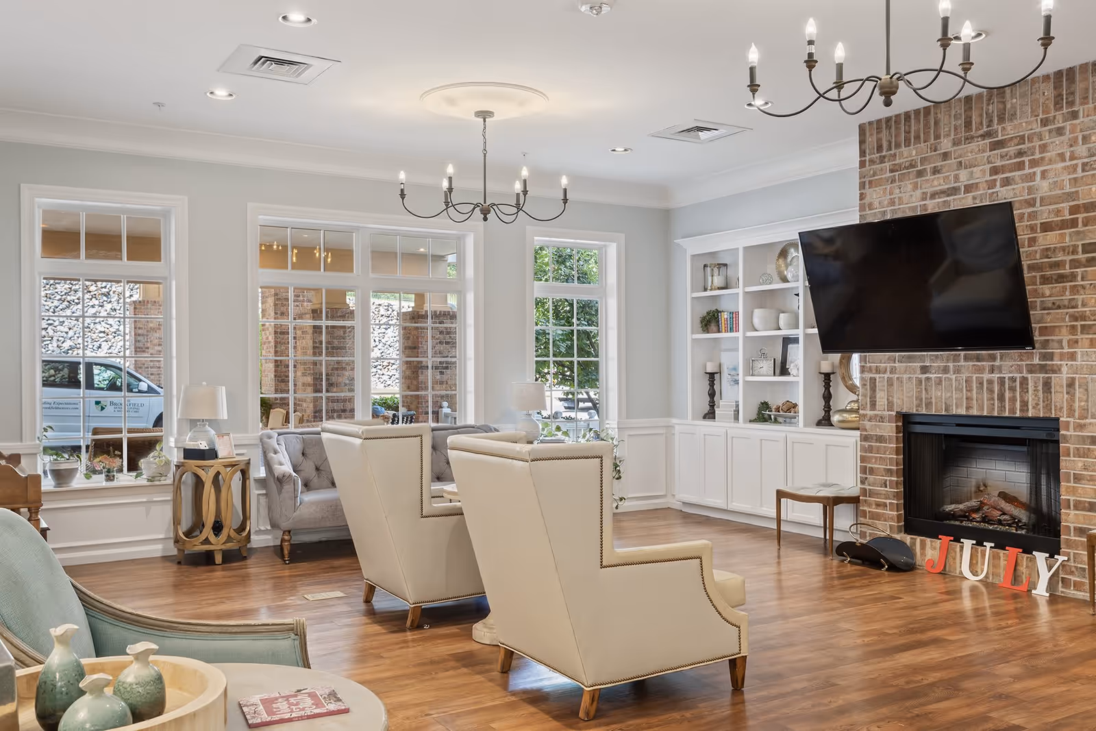 A bright and spacious living room area with large windows, two beige armchairs, a gray tufted sofa, a brick fireplace with a mounted flat-screen TV above it, and built-in white shelves decorated with books, plants, and candles. The floor is wooden, and there are two chandeliers hanging from the ceiling. Outside the windows, a vehicle with Brookfield Assisted Living and Memory Care branding is visible.