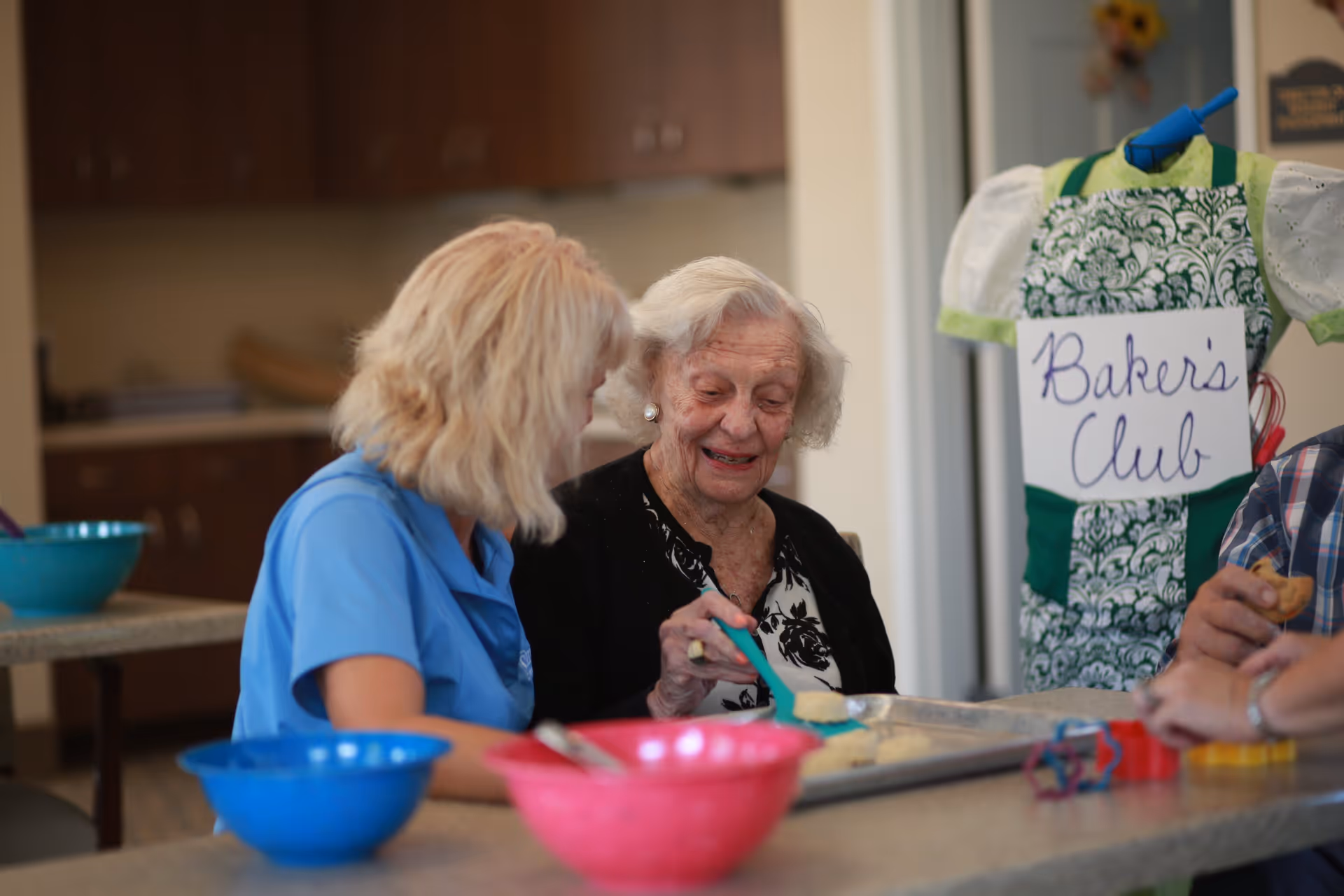 An elderly woman and a caregiver are sitting at a table in a kitchen setting, engaging in a baking activity. The elderly woman is smiling and using a spatula to place dough on a baking tray. Various colorful bowls and cookie cutters are on the table. In the background, there is a mannequin wearing an apron with a sign that reads 'Baker's Club'.