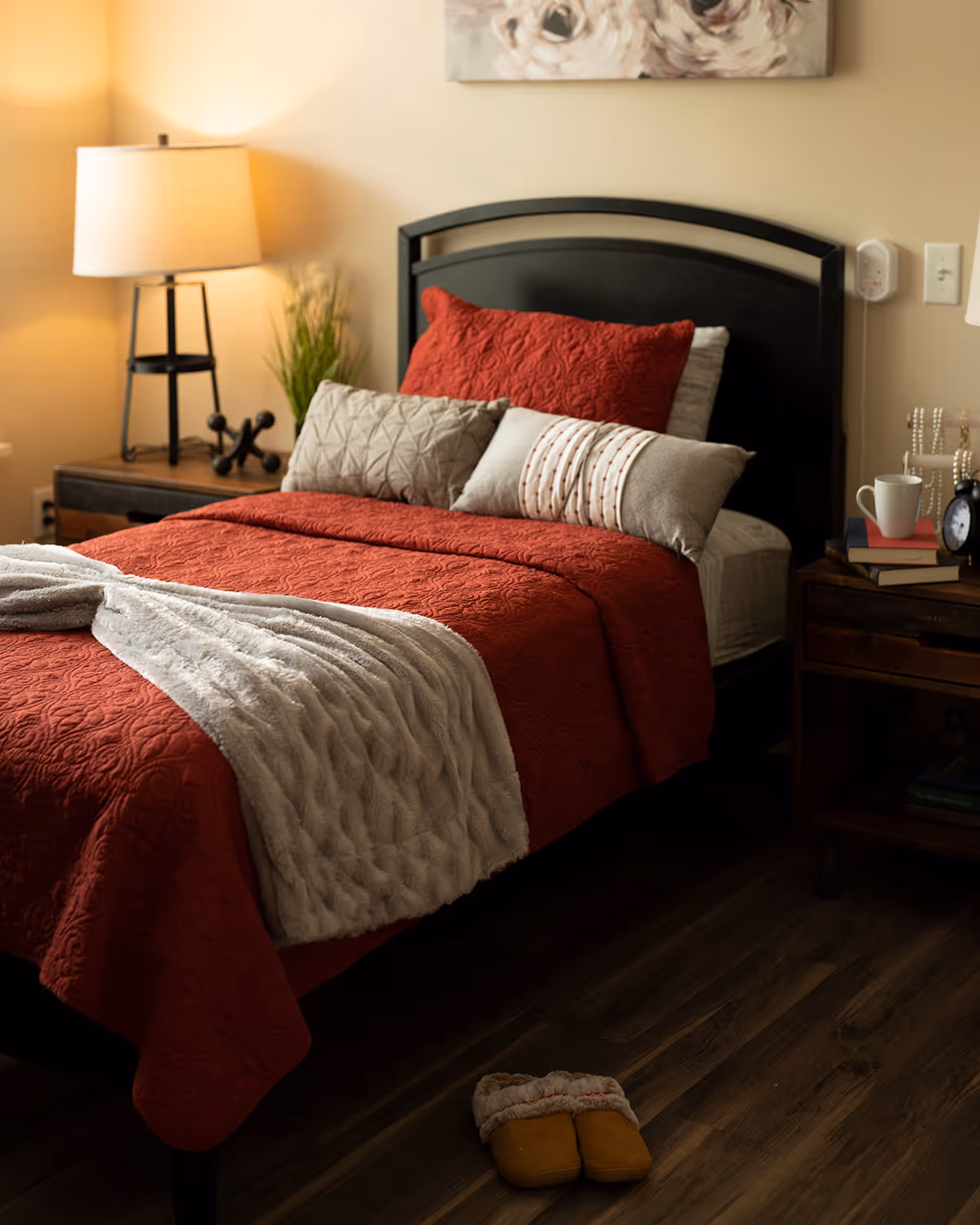 A cozy single bed with a red quilt and decorative pillows, bedside tables with a lamp and books, and slippers on a wooden floor.