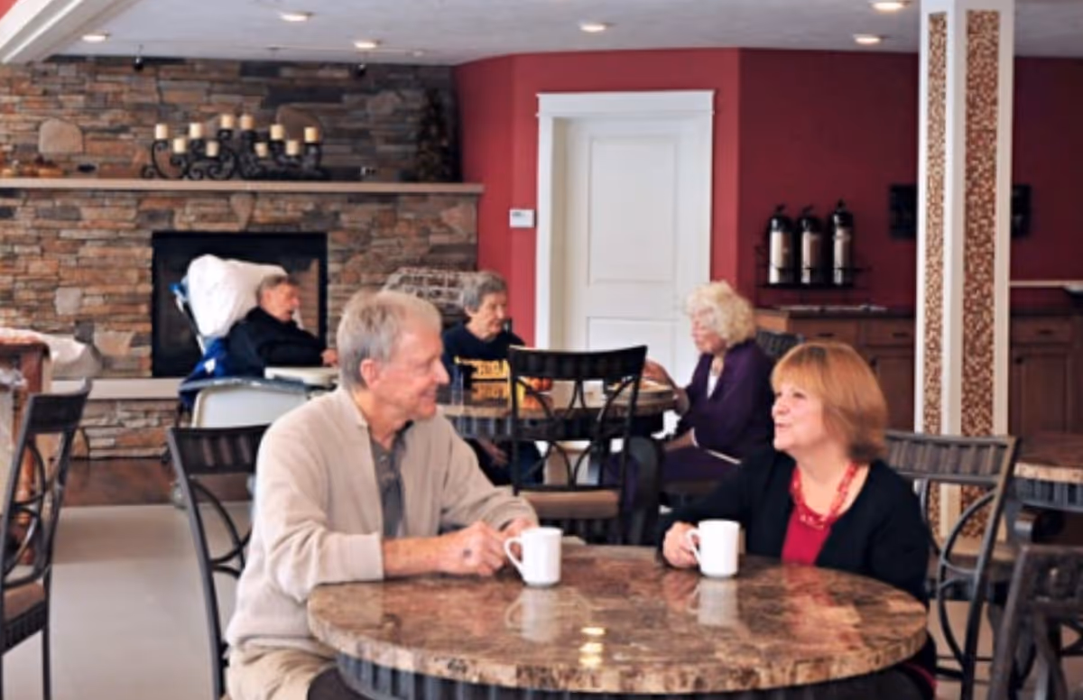 A group of elderly people sitting and conversing at round tables in a cozy room with a stone fireplace and red walls. Two people are seated at the foreground table holding white mugs, while three others are seated at tables in the background.
