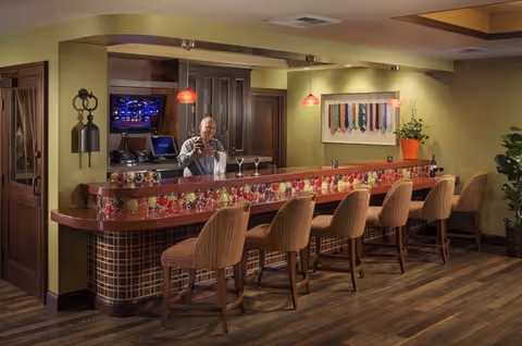 A man pours drinks behind a long tiled bar with six stools in a warm, well-lit lounge.