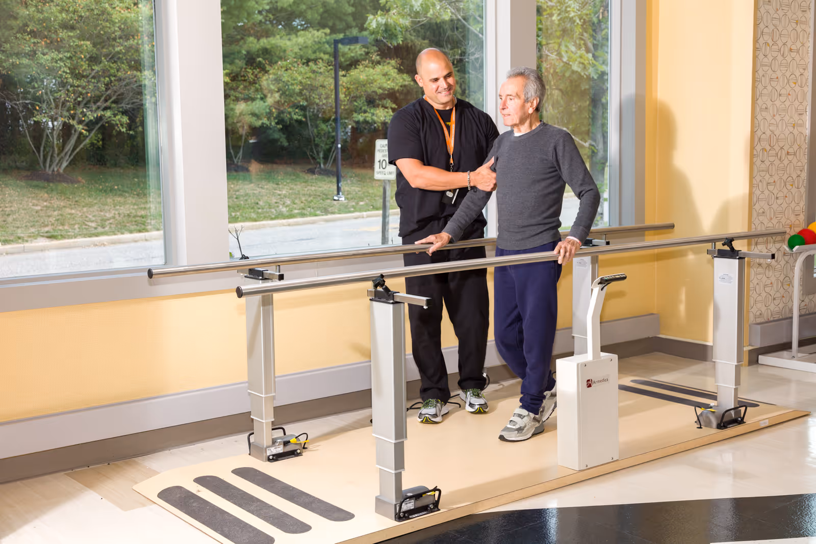 A healthcare worker assists an elderly man as he practices walking with parallel bars in a rehabilitation room with large windows showing greenery outside.