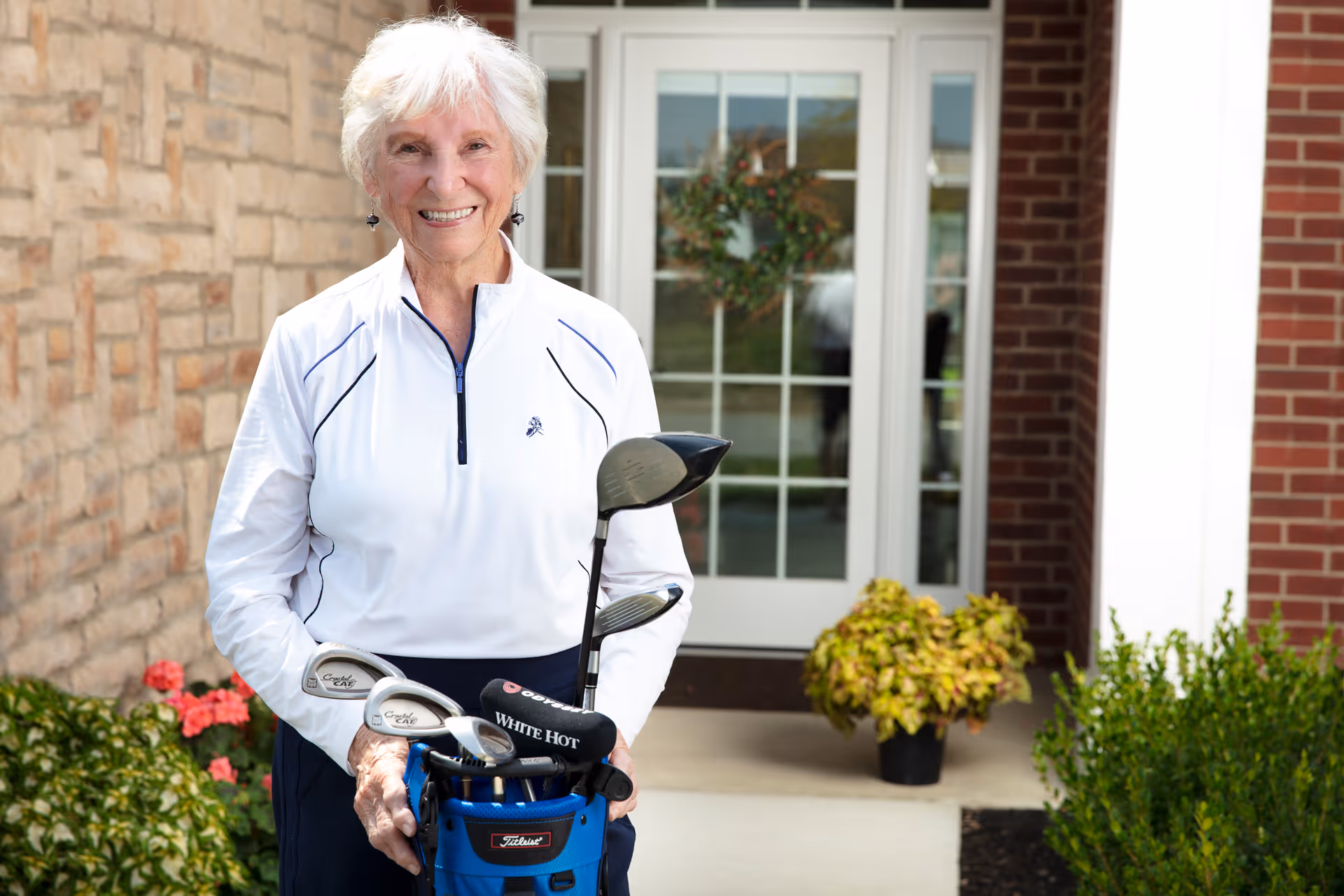 An elderly woman with white hair wearing a white long-sleeve shirt stands outside a building holding a blue golf bag with clubs. Behind her is a glass door with a wreath and some potted plants near the entrance.