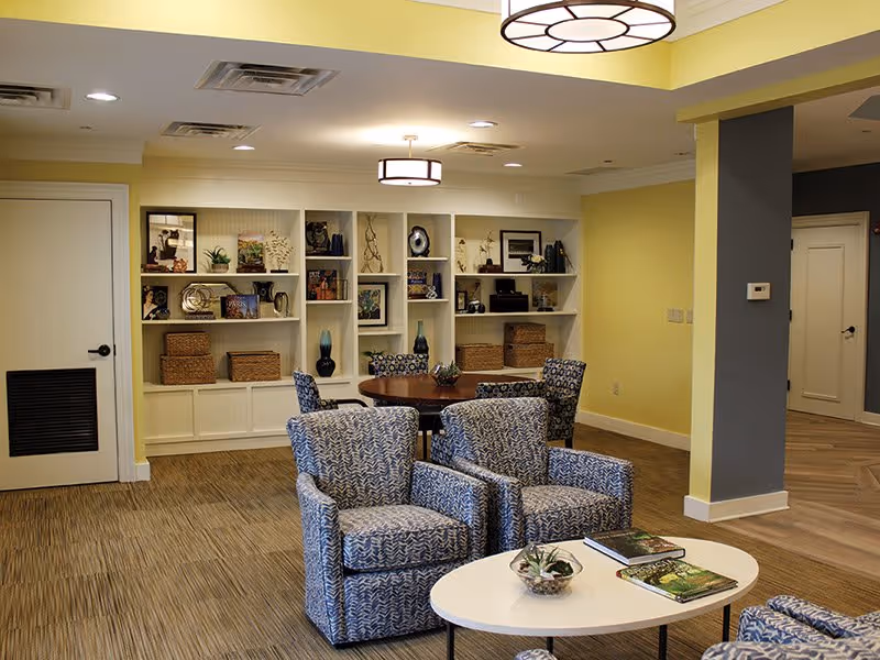A bright communal lounge with patterned armchairs around a coffee table and built-in shelving along a yellow wall.