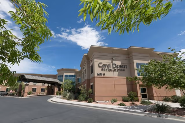 Exterior view of Coral Desert Rehabilitation And Care building under a blue sky with some clouds. The building is two stories with beige and brown walls, surrounded by landscaping with trees and shrubs. A covered driveway entrance is visible on the left side.