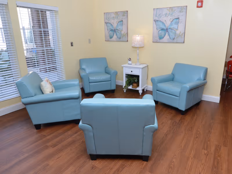 A small sitting area with four light blue armchairs arranged around a white side table and lamp in a room with yellow walls and butterfly artwork.