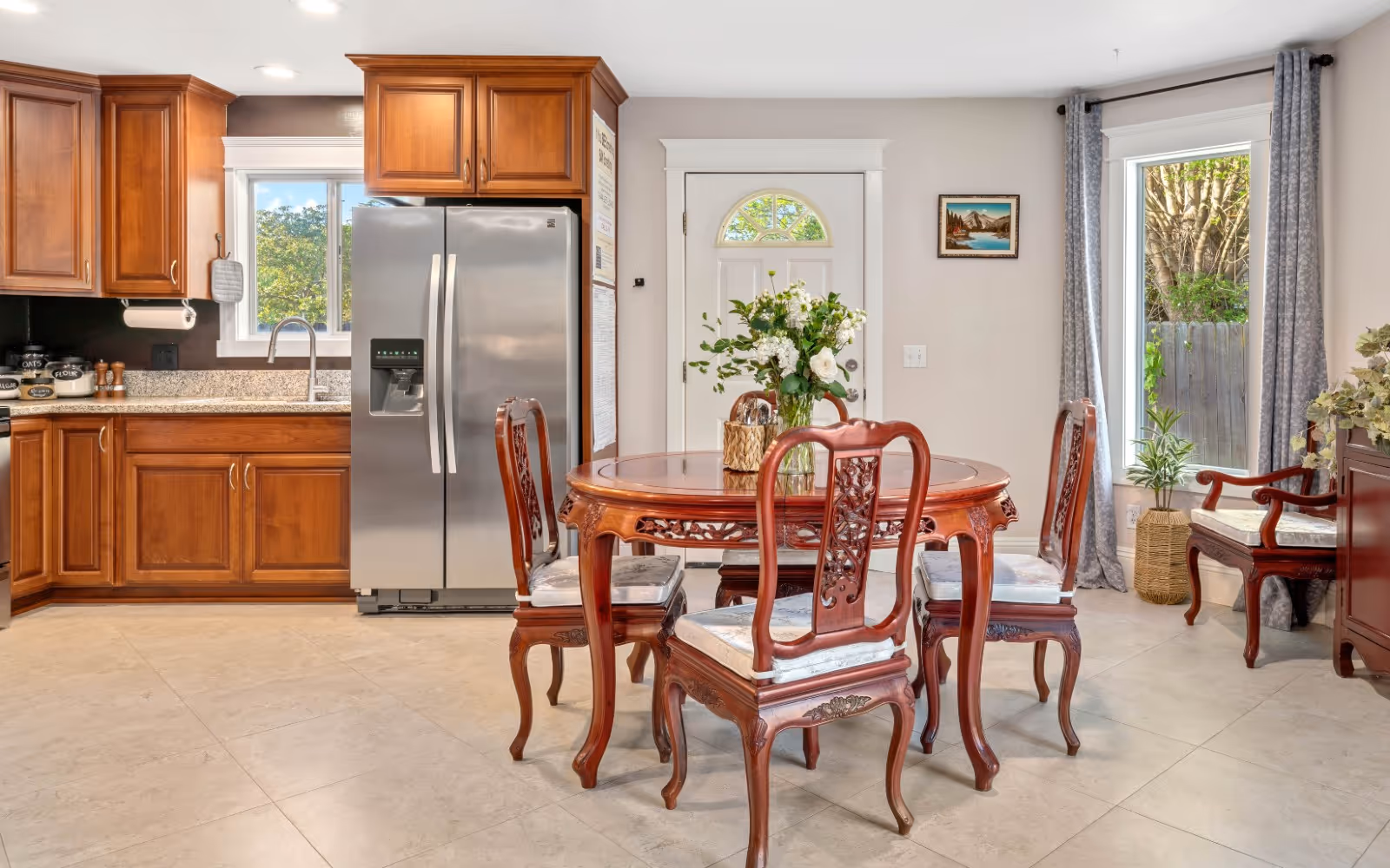 Open kitchen and dining area with wooden cabinets, a stainless steel refrigerator, and a round wooden dining table with chairs and a vase of flowers.