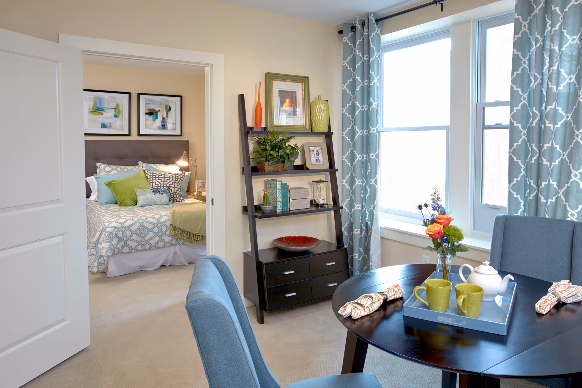 Bright furnished interior showing a dining table with a tray and cups, blue curtains, a decorative shelving unit, and a bedroom visible through an open door.