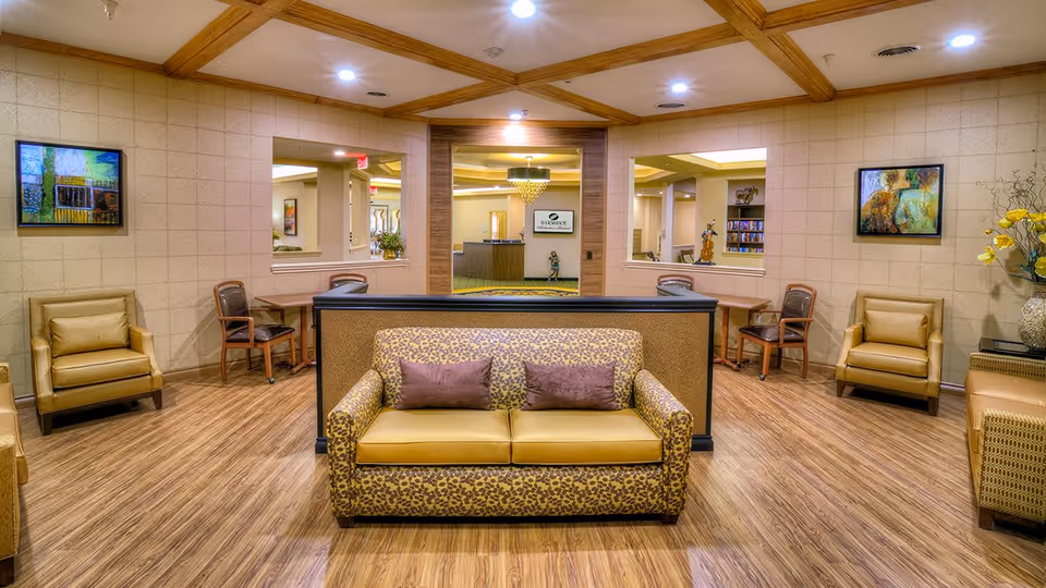 A spacious and well-lit living room area in an assisted living facility featuring a patterned sofa with two purple cushions in the center, flanked by two beige armchairs on each side. The room has wooden flooring, beige walls with framed artwork, and wooden ceiling beams. In the background, there is a reception desk area visible through a large open doorway.