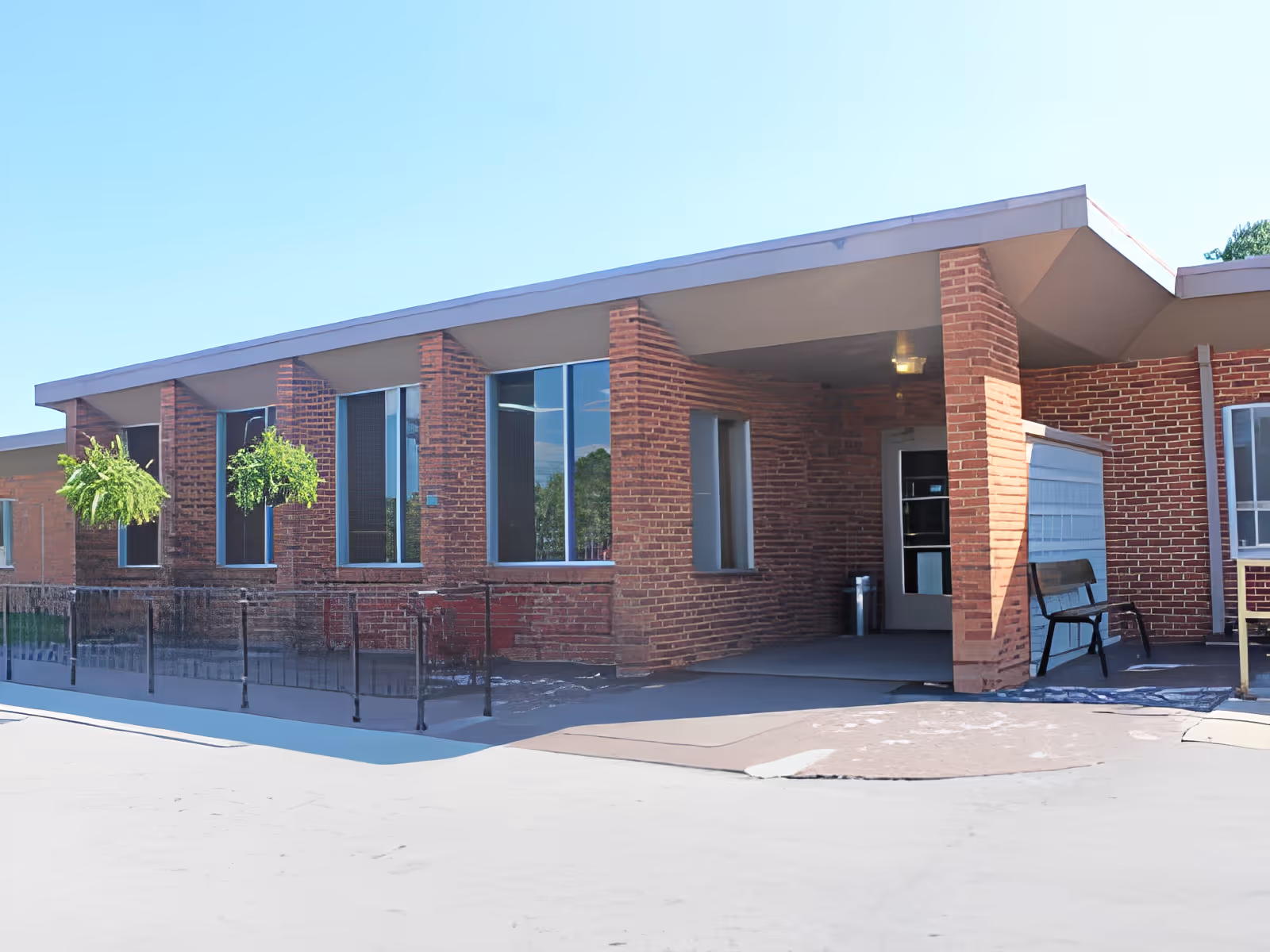 Brick exterior entrance of a single-story building with a covered entry, bench, and hanging plants.