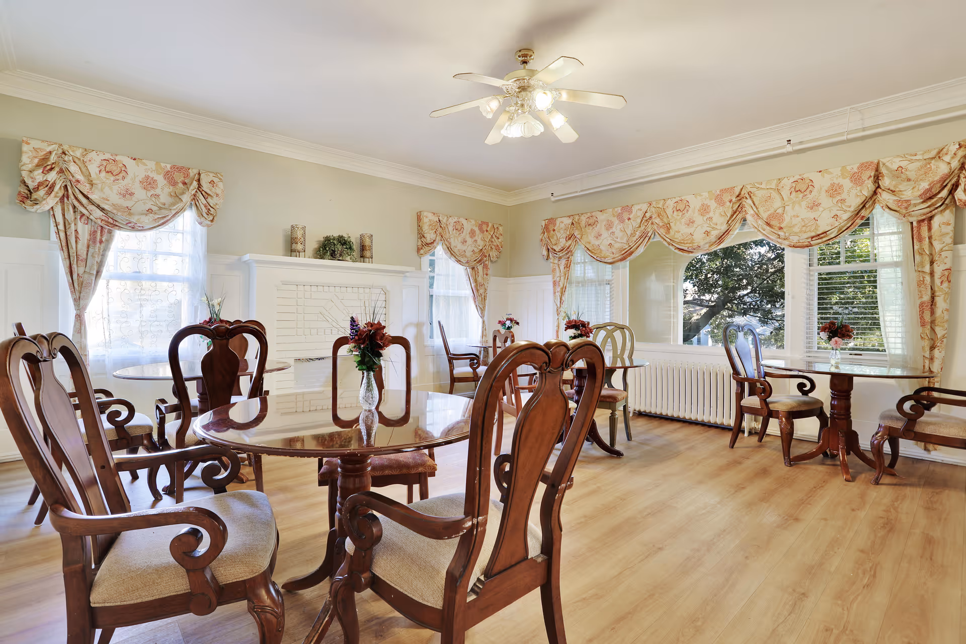 A bright and elegant dining room with wooden tables and chairs arranged neatly. The room features large windows with floral-patterned curtains, a white brick fireplace, and a ceiling fan with lights. Small floral centerpieces are placed on each table, and the floor is covered with light wood laminate.