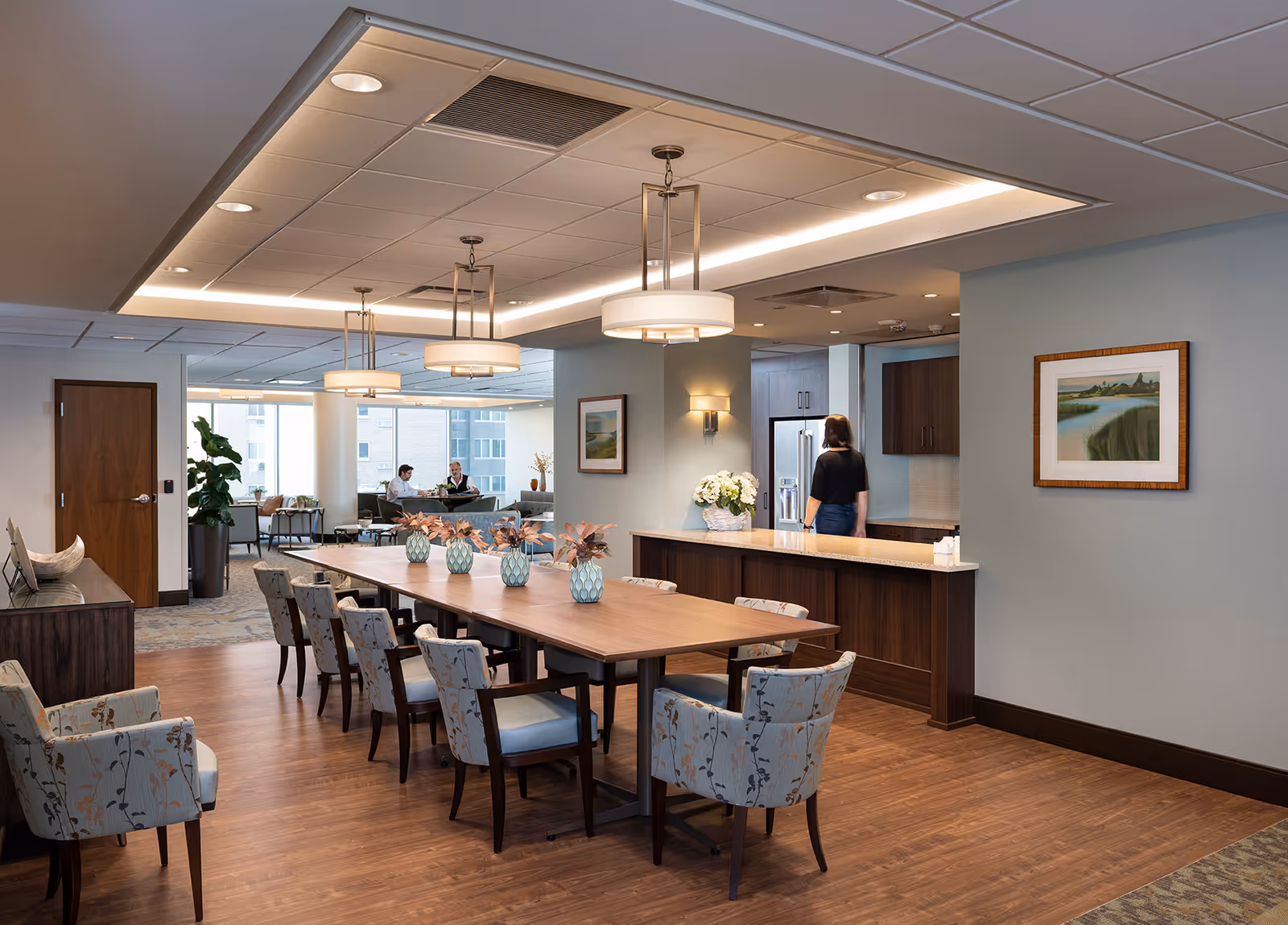 A spacious, well-lit communal dining area with a long wooden table surrounded by upholstered chairs, pendant lights, and a kitchenette where a person stands at the counter.