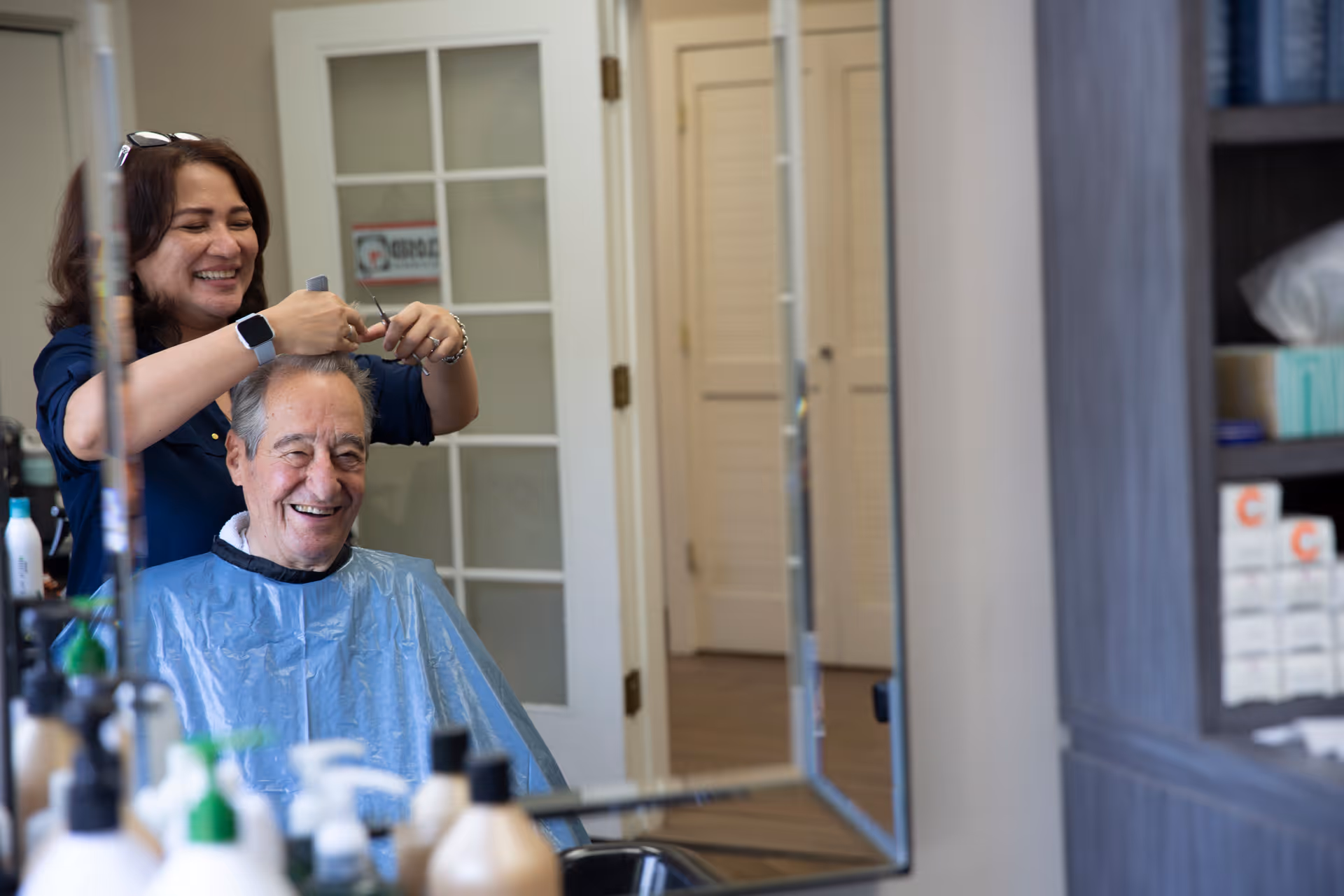 A smiling elderly man wearing a blue haircut cape is getting his hair cut by a woman in a salon setting. The woman is also smiling and holding scissors near the man's hair. The scene is reflected in a mirror, with various hair care products visible in the foreground and shelves with supplies in the background.